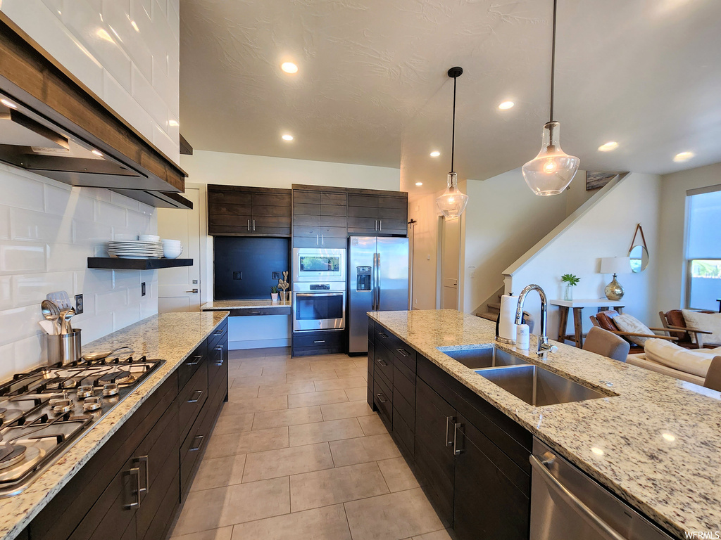 Kitchen featuring backsplash, appliances with stainless steel finishes, dark brown cabinetry, light stone countertops, pendant lighting, and light tile flooring