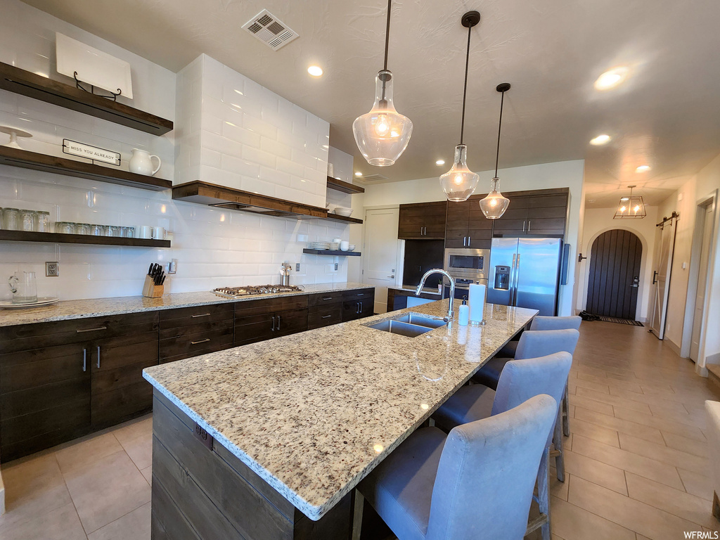 Kitchen with kitchen island with sink, backsplash, light tile floors, appliances with stainless steel finishes, a center island, dark brown cabinetry, light stone countertops, and pendant lighting