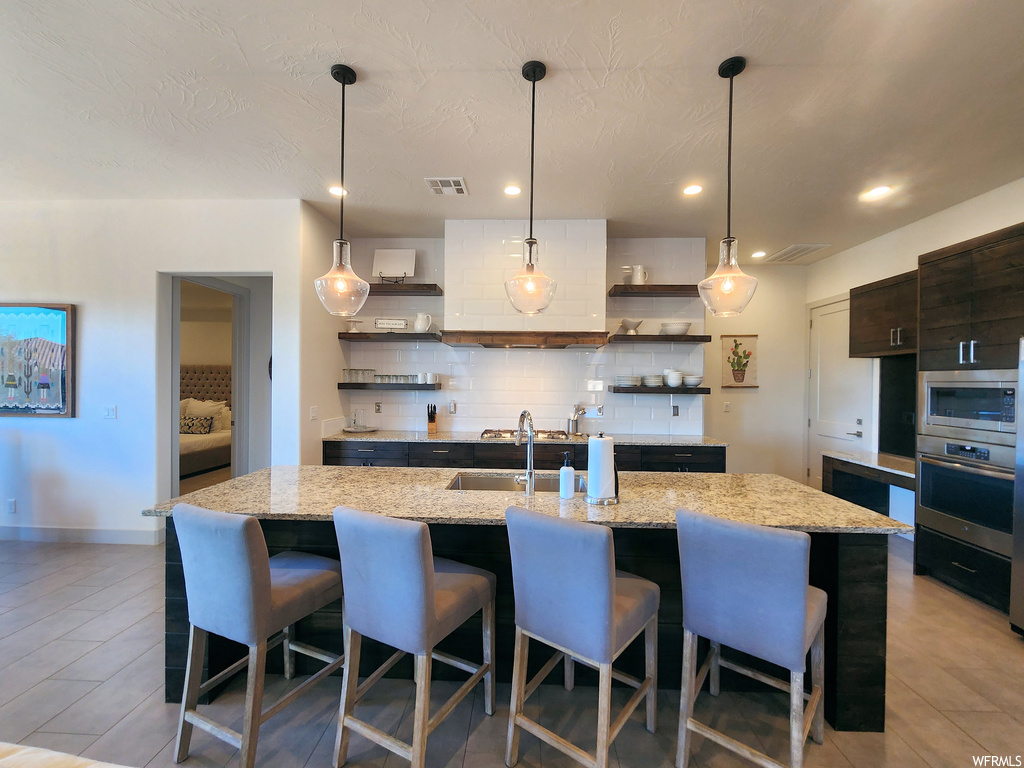 Kitchen featuring hanging light fixtures, backsplash, a kitchen island, light stone countertops, wall chimney exhaust hood, and stainless steel microwave
