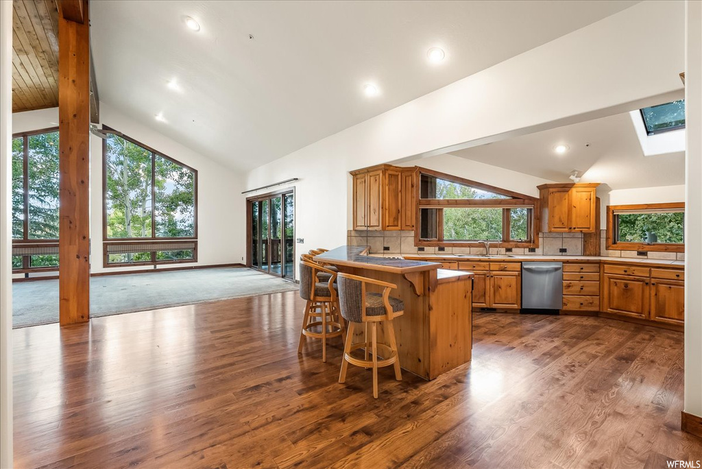 Kitchen featuring stainless steel dishwasher, backsplash, brown cabinets, light countertops, light hardwood floors, a center island, and vaulted ceiling with skylight