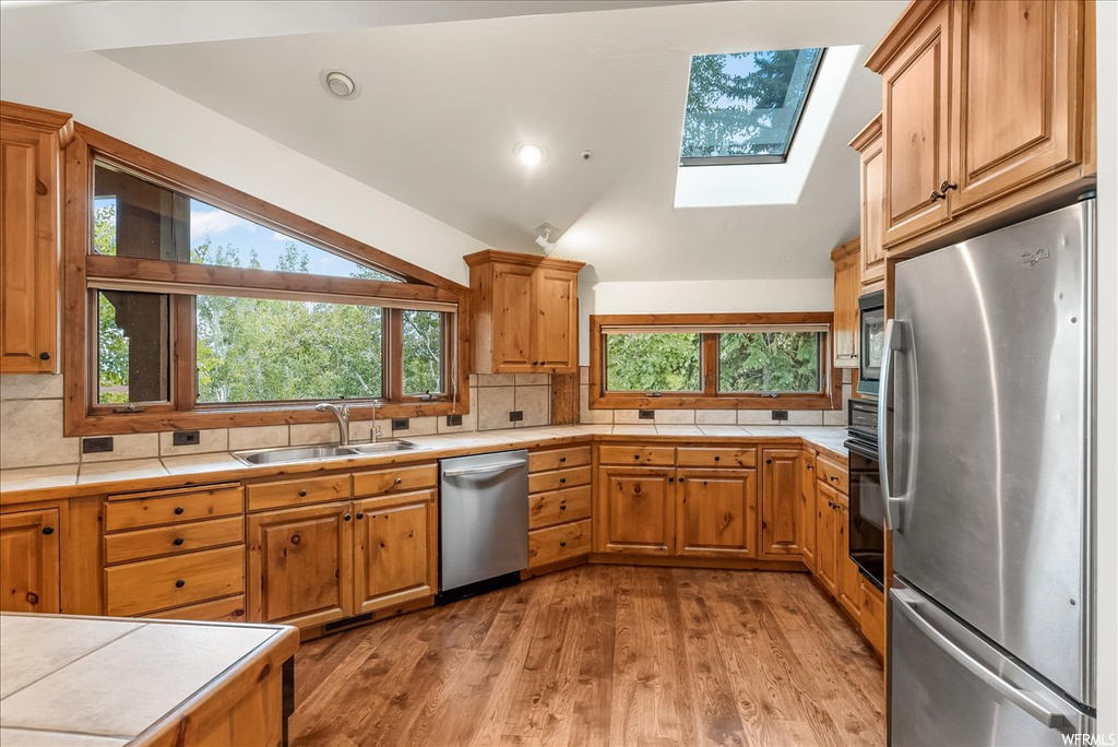 Kitchen with appliances with stainless steel finishes, plenty of natural light, lofted ceiling with skylight, brown cabinets, light countertops, light hardwood flooring, and backsplash