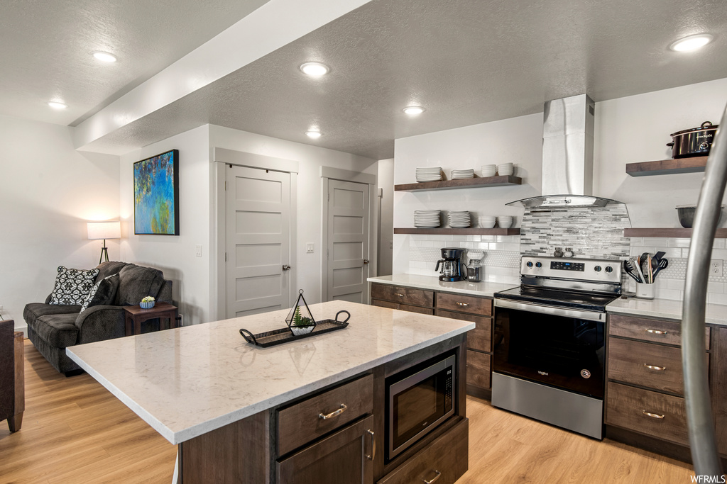 Kitchen featuring backsplash, wall chimney range hood, light hardwood flooring, dark brown cabinetry, a kitchen island with sink, and stainless steel appliances