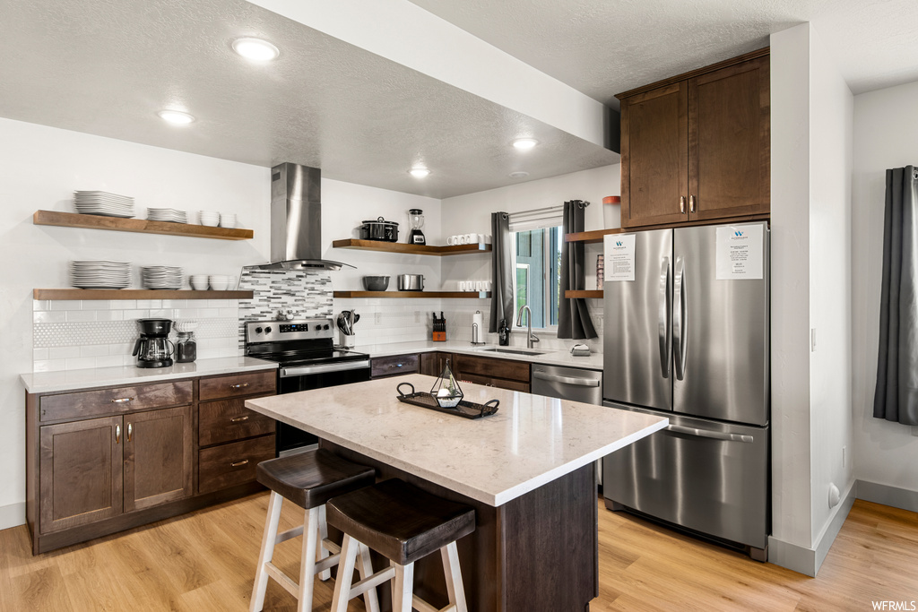 Kitchen with stainless steel appliances, a textured ceiling, a center island, range hood, an island with sink, light stone counters, dark brown cabinets, light hardwood flooring, and backsplash