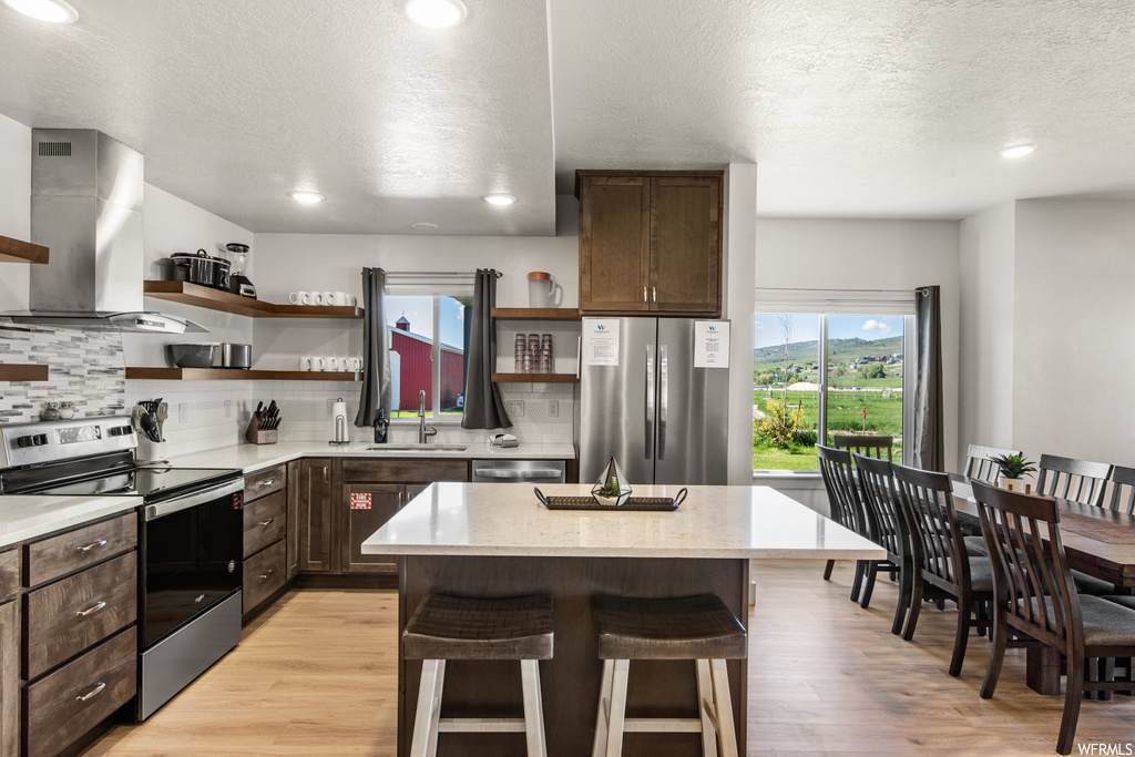 Kitchen featuring wall chimney range hood, a kitchen island, stainless steel appliances, a textured ceiling, dark brown cabinetry, light countertops, backsplash, and light hardwood floors