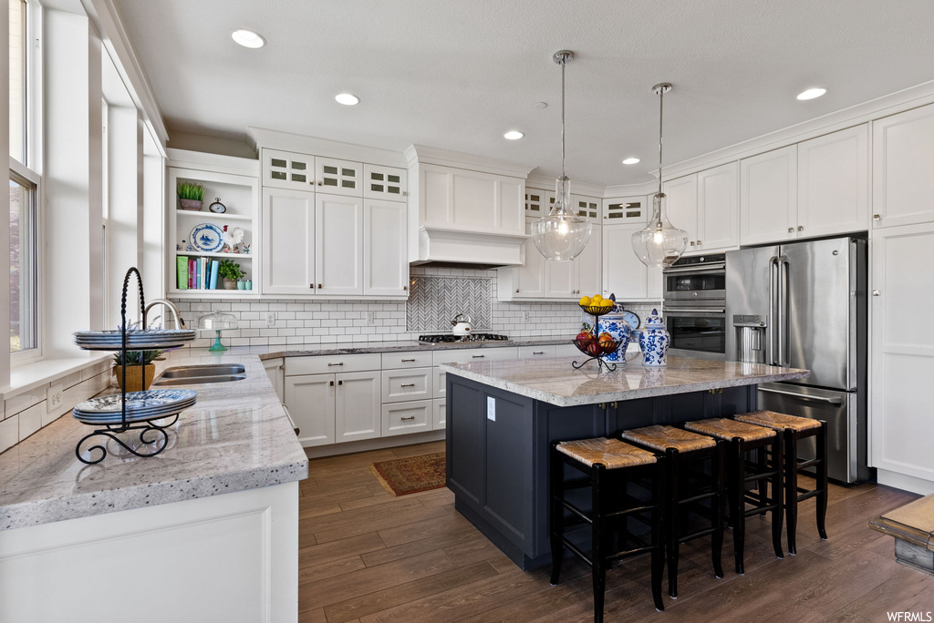 Kitchen with decorative light fixtures, kitchen island with sink, backsplash, light countertops, custom exhaust hood, white cabinetry, appliances with stainless steel finishes, a center island, and hardwood floors