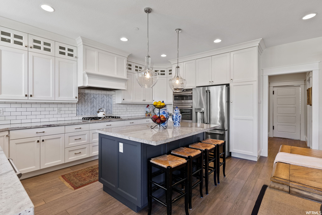 Kitchen with appliances with stainless steel finishes, custom range hood, a center island, an island with sink, white cabinets, light countertops, hanging light fixtures, backsplash, and light hardwood floors