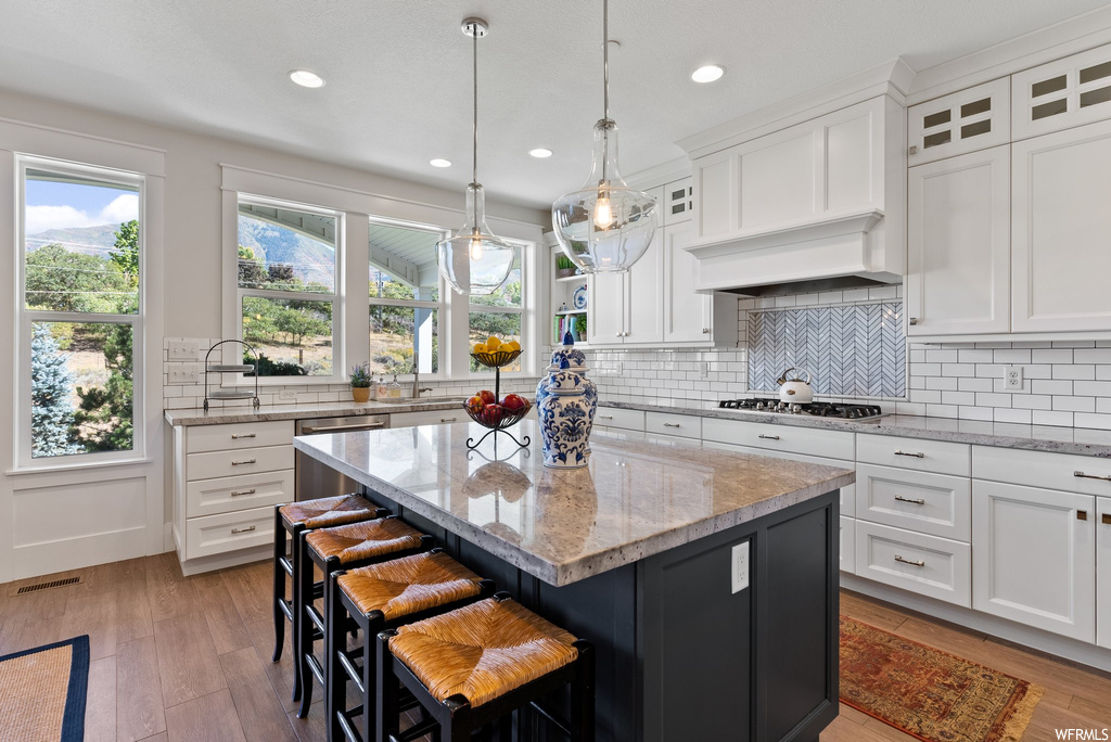 Kitchen with a kitchen island, white cabinetry, custom range hood, pendant lighting, stainless steel appliances, light stone countertops, hardwood flooring, a center island with sink, and backsplash