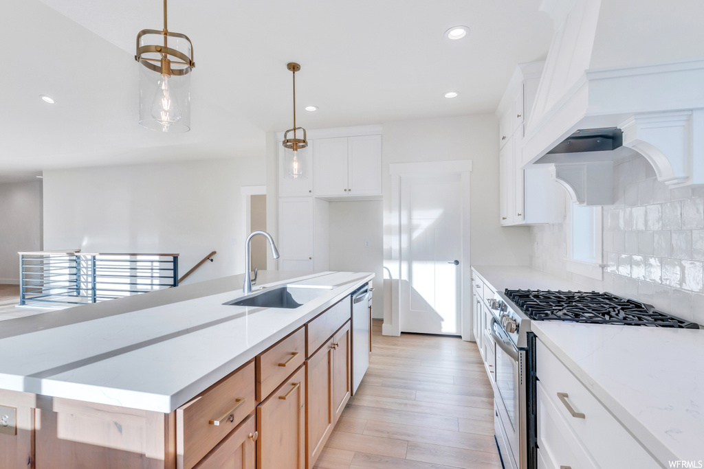 Kitchen featuring backsplash, white cabinets, light countertops, premium range hood, light hardwood flooring, pendant lighting, and stainless steel appliances