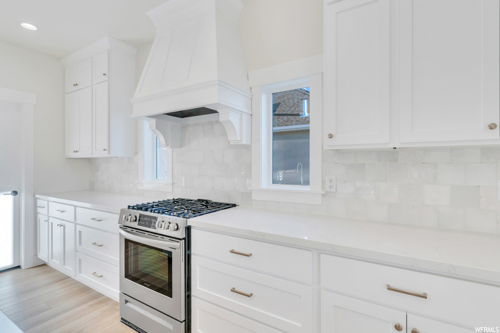 Kitchen featuring white cabinets, stainless steel range with gas stovetop, light countertops, premium range hood, backsplash, and light hardwood floors