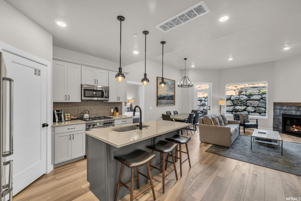 Kitchen featuring hanging light fixtures, kitchen island with sink, backsplash, white cabinets, light countertops, a kitchen island, light hardwood flooring, stainless steel appliances, and a fireplace