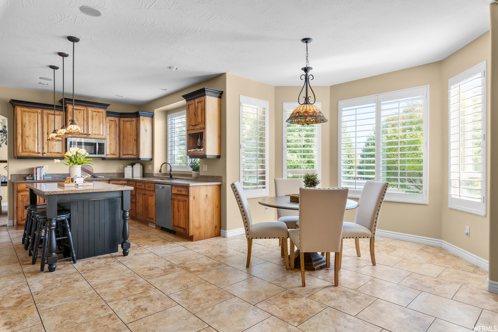 Kitchen featuring appliances with stainless steel finishes, pendant lighting, light tile flooring, brown cabinets, light countertops, and a healthy amount of sunlight