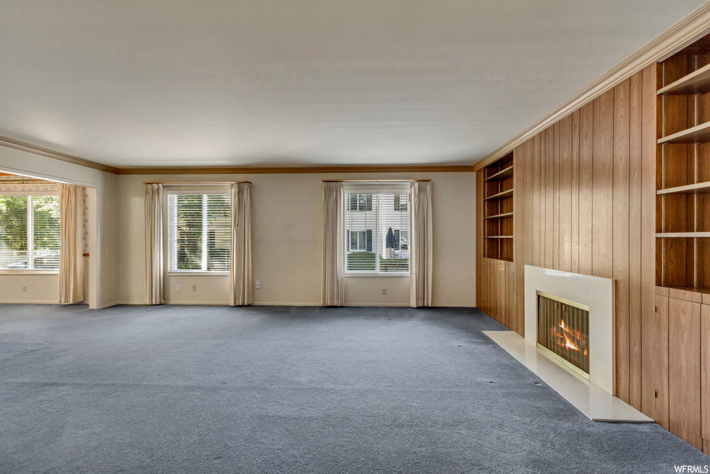 Carpeted living room with plenty of natural light, built in shelves, crown molding, wooden walls, and a fireplace