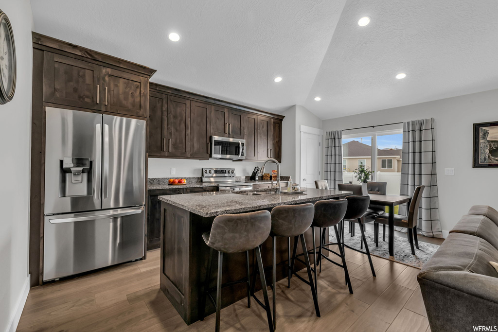 Kitchen featuring lofted ceiling, a kitchen island with sink, light hardwood flooring, appliances with stainless steel finishes, and dark stone counters