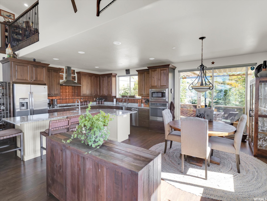 Kitchen featuring dark hardwood flooring, wall chimney range hood, a center island, stainless steel appliances, and decorative light fixtures