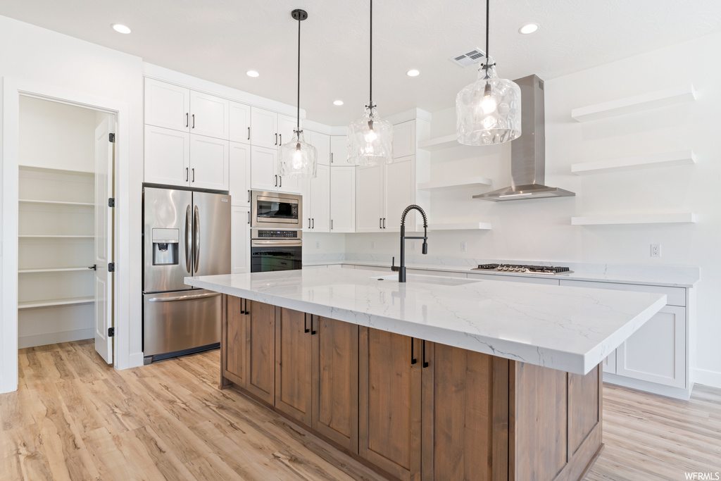 Kitchen featuring wall chimney range hood, light hardwood flooring, white cabinetry, hanging light fixtures, and an island with sink