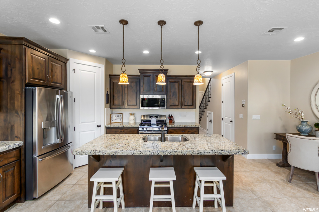 Kitchen with light stone countertops, a kitchen island with sink, light tile floors, appliances with stainless steel finishes, and hanging light fixtures
