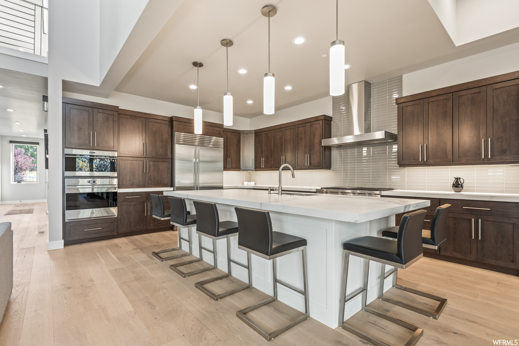 Kitchen featuring backsplash, light countertops, light hardwood floors, a center island, dark brown cabinetry, stainless steel appliances, pendant lighting, a kitchen island with sink, and wall chimney exhaust hood