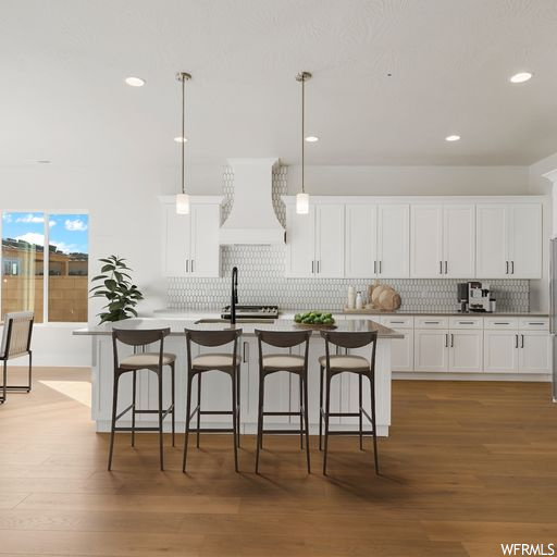 Kitchen featuring white cabinets, custom exhaust hood, light hardwood floors, hanging light fixtures, and a kitchen island with sink