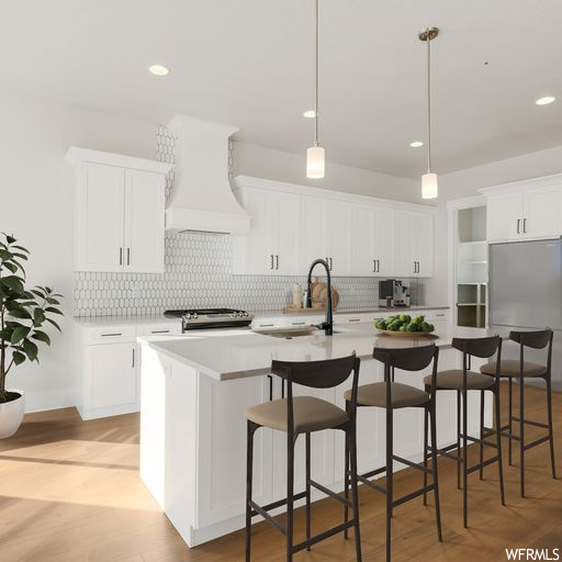 Kitchen with decorative light fixtures, white cabinetry, custom range hood, and a kitchen island with sink