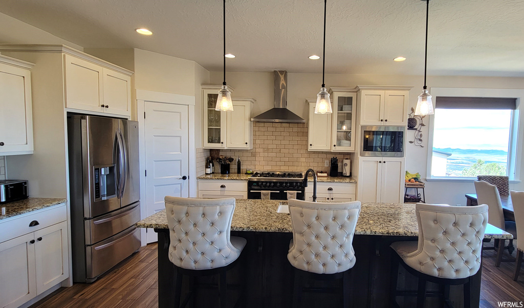 Kitchen featuring pendant lighting, a center island with sink, a kitchen bar, appliances with stainless steel finishes, and wall chimney exhaust hood