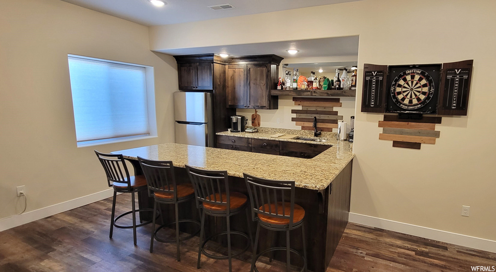 Kitchen with dark brown cabinetry, stainless steel fridge, light stone countertops, dark hardwood floors, and a kitchen breakfast bar