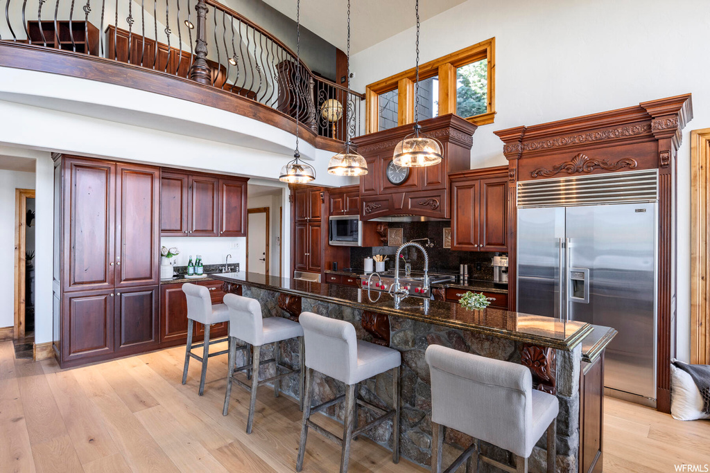 Kitchen with a kitchen island with sink, built in appliances, a towering ceiling, light hardwood / wood-style flooring, and a kitchen breakfast bar