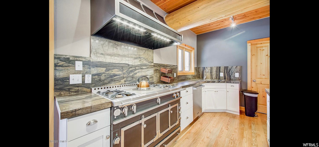 Kitchen with light hardwood flooring, wall chimney exhaust hood, backsplash, dishwasher, and white cabinetry