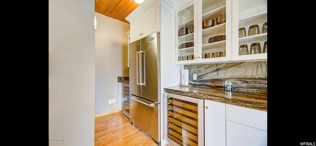 Bar featuring high end refrigerator, light hardwood flooring, white cabinets, backsplash, and dark stone counters