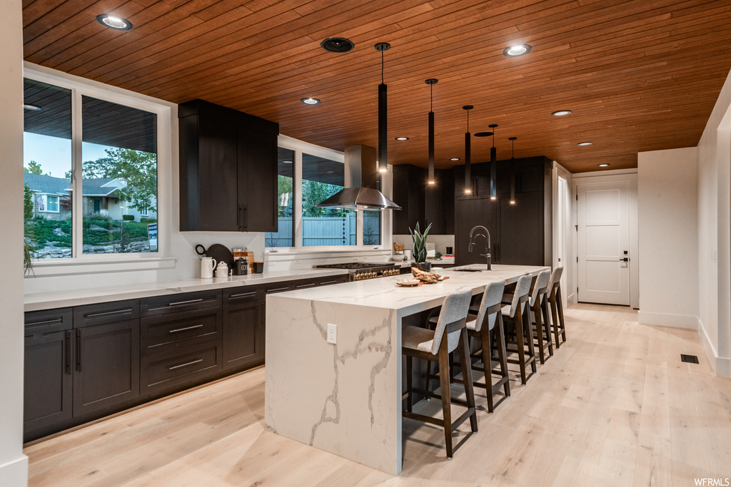 Kitchen featuring decorative light fixtures, light hardwood floors, a kitchen island with sink, ventilation hood, and light stone counters