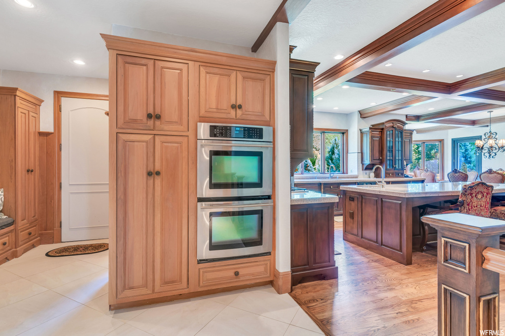 Kitchen featuring a center island with sink, double oven, light hardwood flooring, a notable chandelier, and beam ceiling