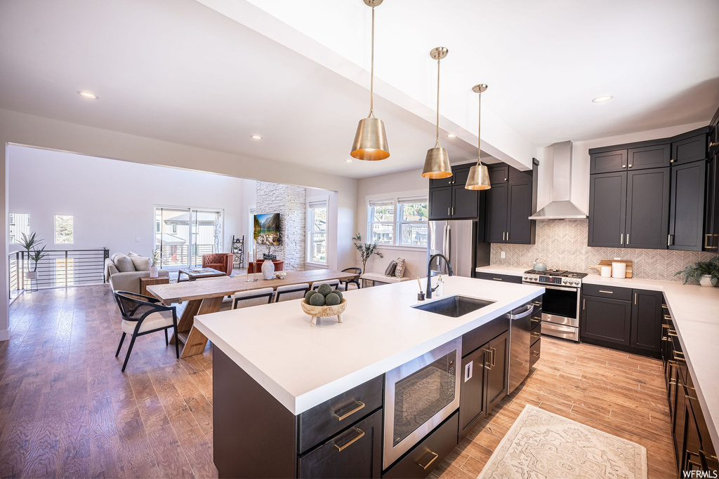 Kitchen featuring pendant lighting, light hardwood flooring, appliances with stainless steel finishes, wall chimney range hood, and an island with sink