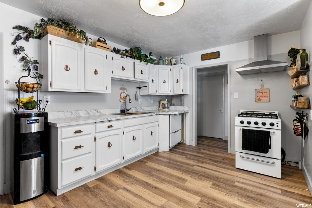 Kitchen with white cabinets, white range with gas stovetop, wall chimney exhaust hood, and light hardwood flooring