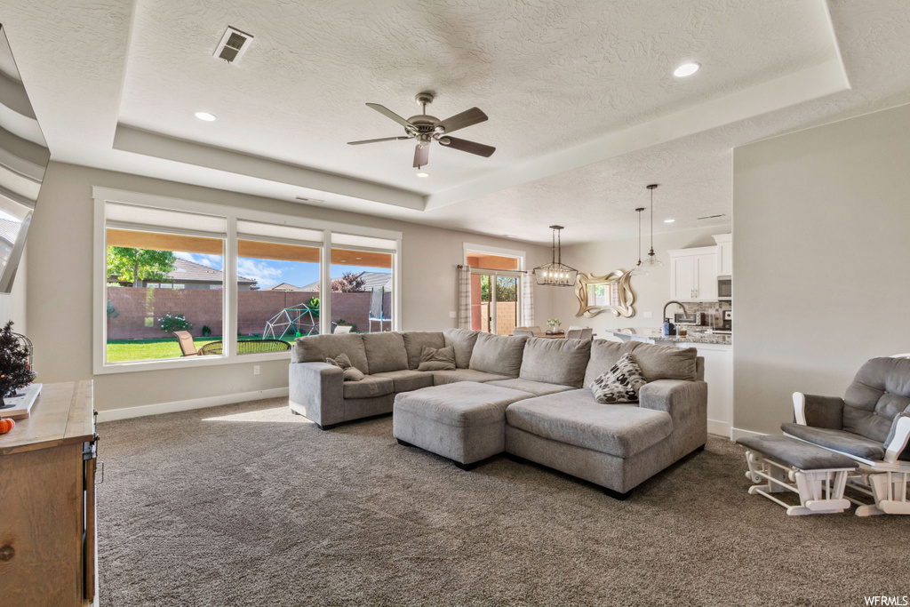 Carpeted living room with a raised ceiling, a healthy amount of sunlight, and ceiling fan with notable chandelier