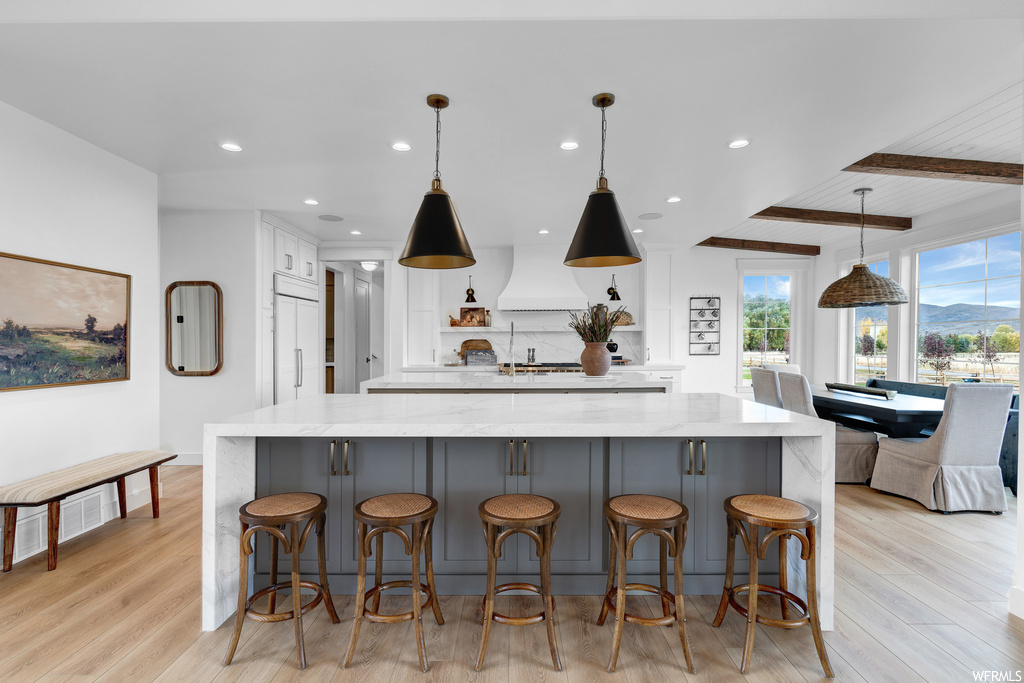 Kitchen featuring white cabinets, light hardwood floors, premium range hood, beam ceiling, and decorative light fixtures