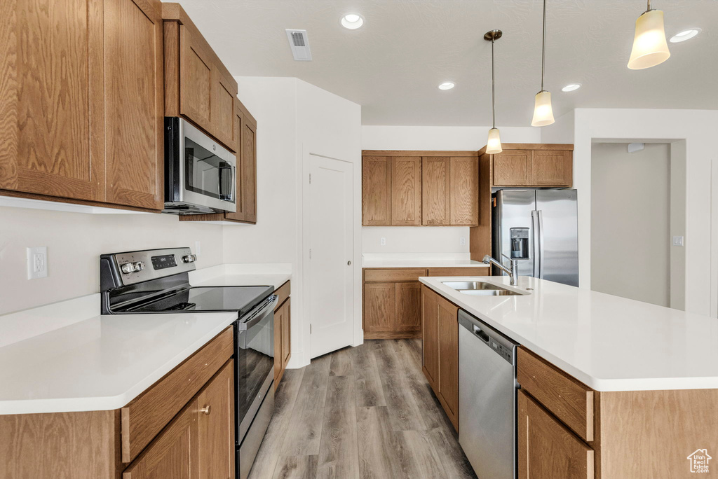 Kitchen with appliances with stainless steel finishes, light hardwood / wood-style floors, an island with sink, sink, and decorative light fixtures