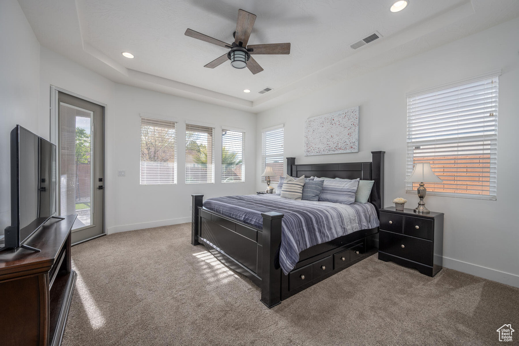 Carpeted bedroom featuring a raised ceiling, multiple windows, ceiling fan, and access to exterior