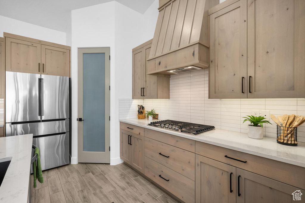 Kitchen featuring tasteful backsplash, light wood-type flooring, stainless steel appliances, premium range hood, and light stone countertops