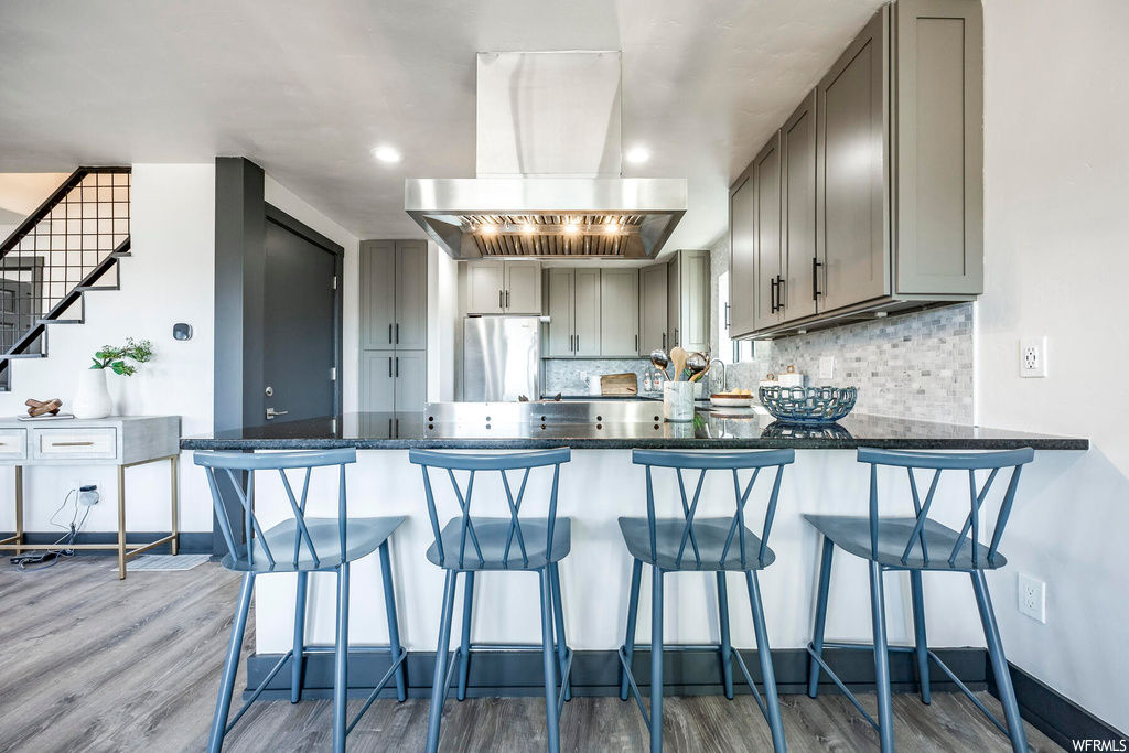 Kitchen featuring island exhaust hood, light hardwood / wood-style floors, a breakfast bar, gray cabinets, and backsplash