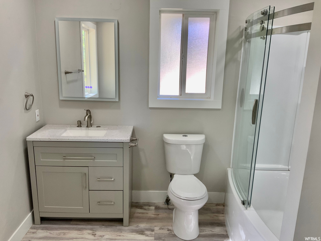 Full bathroom with hardwood flooring, large vanity, combined bath / shower with glass door, and toilet
