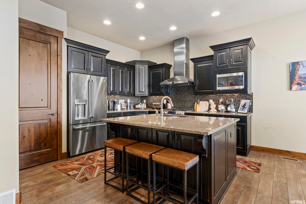 Kitchen with wall chimney range hood, backsplash, light hardwood floors, a kitchen island with sink, and appliances with stainless steel finishes