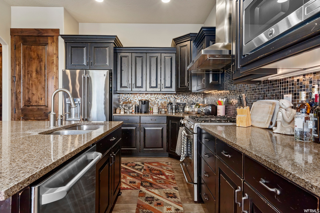 Kitchen with sink, wall chimney range hood, backsplash, premium appliances, and light stone countertops