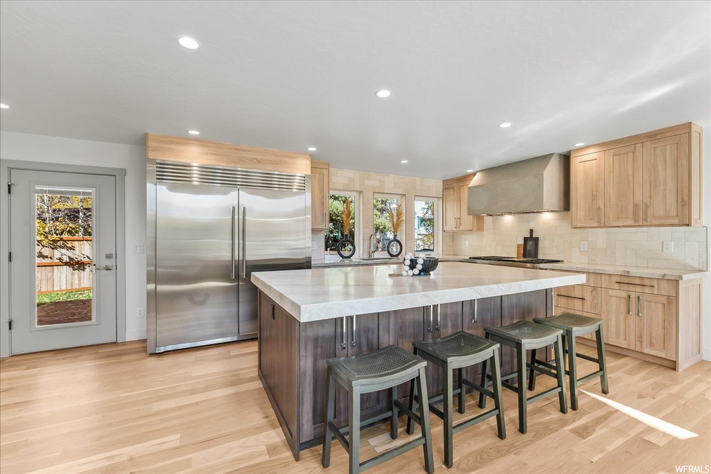 Kitchen featuring wall chimney exhaust hood, a center island, light hardwood / wood-style floors, backsplash, and stainless steel appliances