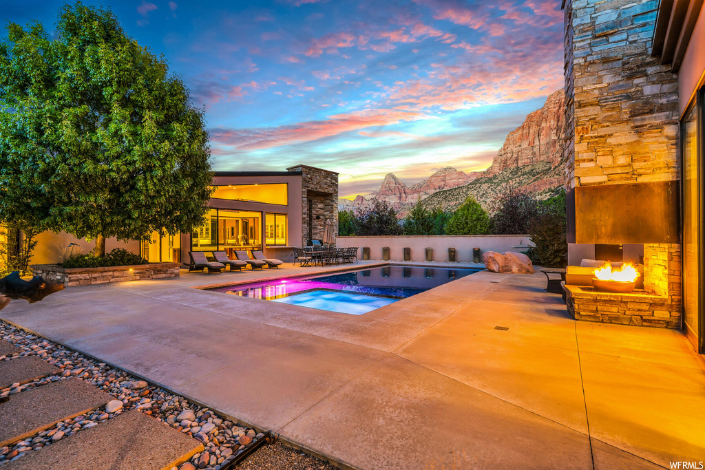 Pool at dusk with an outdoor stone fireplace, a mountain view, and a patio