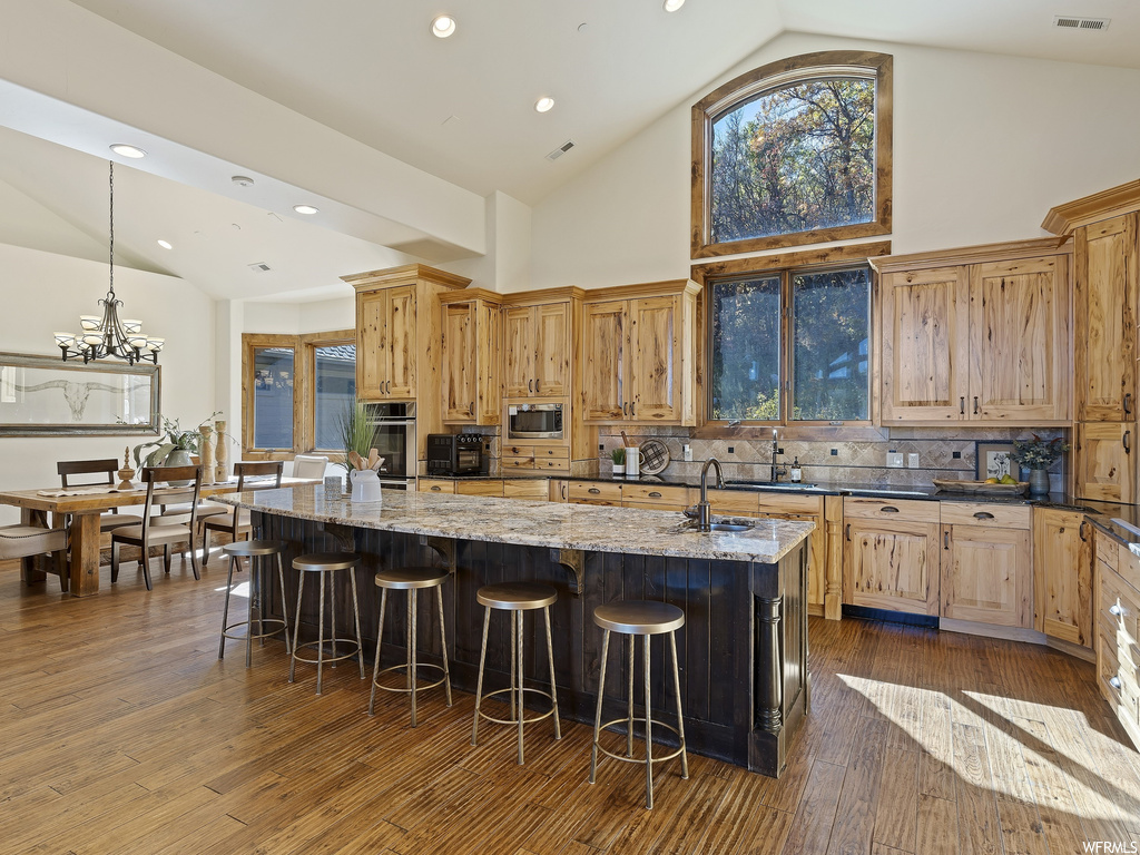 Kitchen featuring decorative light fixtures, dark hardwood / wood-style floors, high vaulted ceiling, appliances with stainless steel finishes, and a kitchen island with sink
