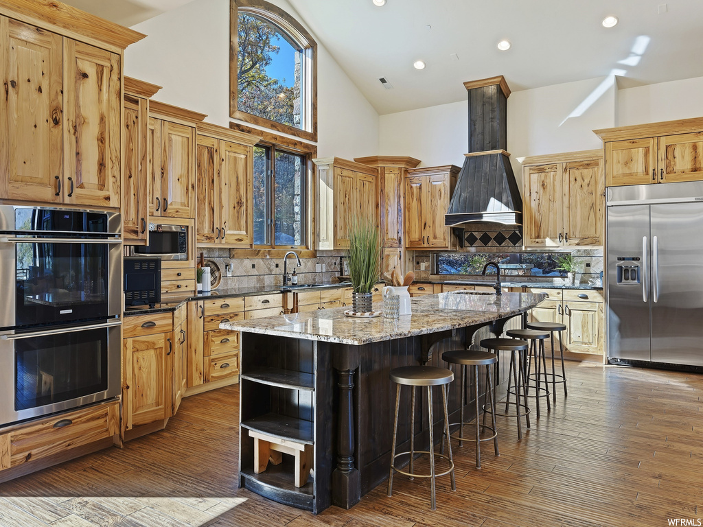 Kitchen with hardwood / wood-style flooring, a center island with sink, high vaulted ceiling, light stone countertops, and backsplash