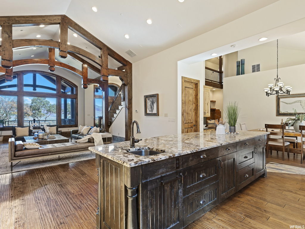 Kitchen with light stone countertops, light hardwood / wood-style floors, high vaulted ceiling, a center island with sink, and a notable chandelier