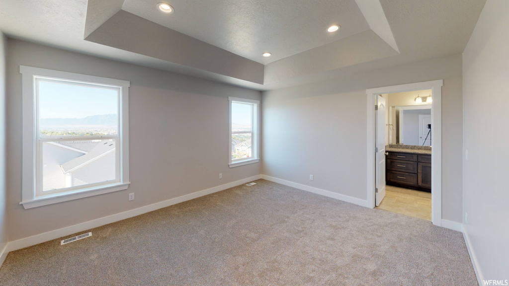 Unfurnished bedroom with light colored carpet, ensuite bathroom, and a tray ceiling