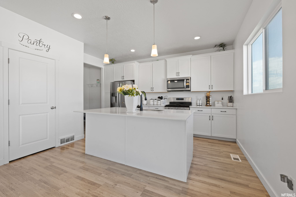 Kitchen featuring a center island with sink, light hardwood / wood-style flooring, stainless steel appliances, hanging light fixtures, and white cabinets