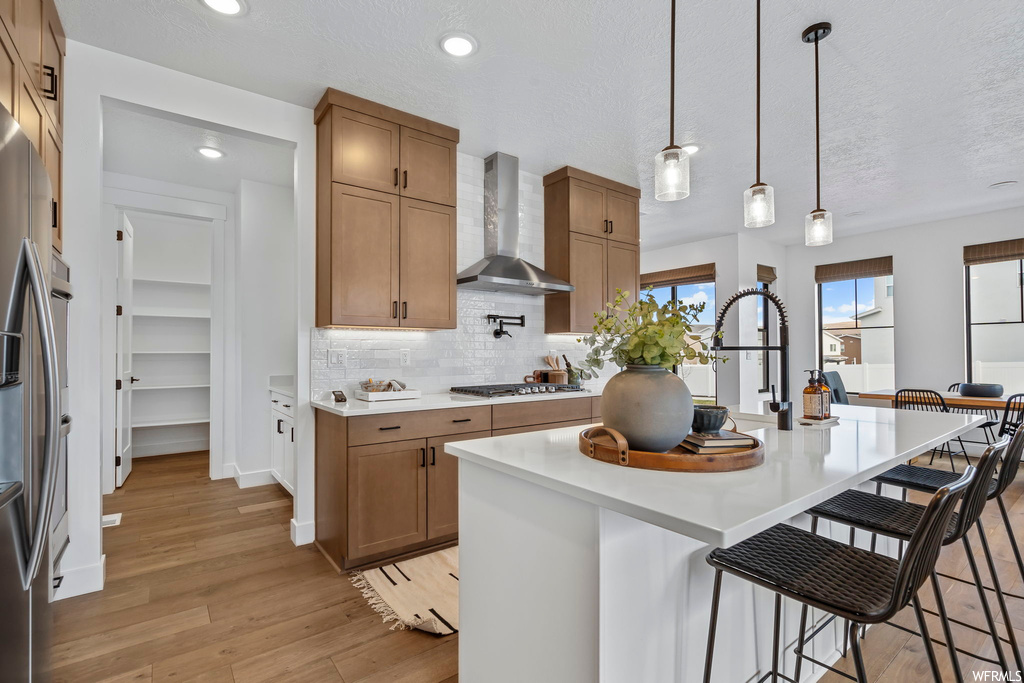 Kitchen featuring light hardwood / wood-style flooring, hanging light fixtures, a breakfast bar area, a center island with sink, and wall chimney range hood
