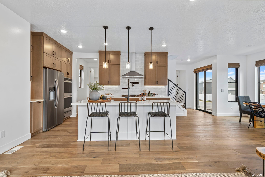 Kitchen featuring light hardwood / wood-style flooring, stainless steel fridge with ice dispenser, hanging light fixtures, backsplash, and wall chimney range hood
